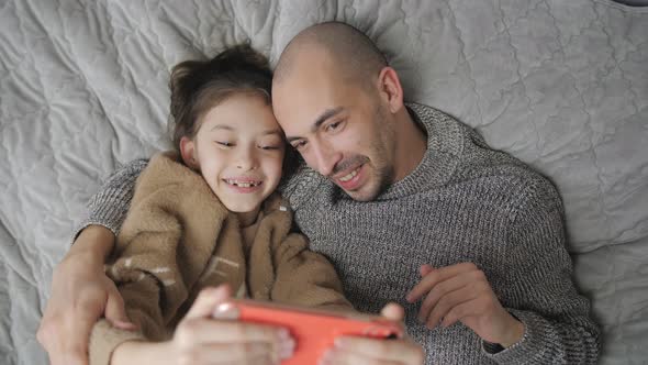 Father and Daughter Lie on the Bed and Communicate Via Video Link on a Smartphone