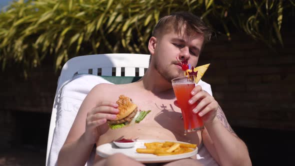 Young Bearded Man Drinking a Cocktail From a Plastic Tube and Eats Hamburger While Relaxing on