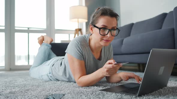 Woman with Glasses is Lying on the Floor and Makes an Online Purchase Using a Credit Card and Laptop alt