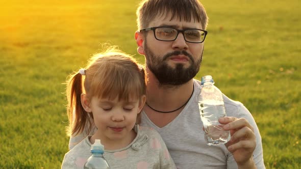 Happy Family Sit on Green Lawn at Sunset Look at Camera and Drink Water From Bottle in Summer Park