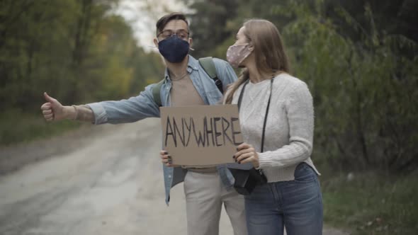 Young Caucasian Boyfriend and Girlfriend in Covid19 Face Masks Hitchhiking with Anywhere Cardboard alt
