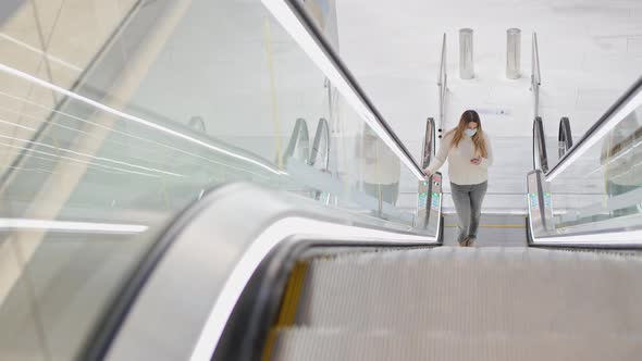 a Young Woman in a Protective Mask Climbs the Escalator and Looks at the Phone alt