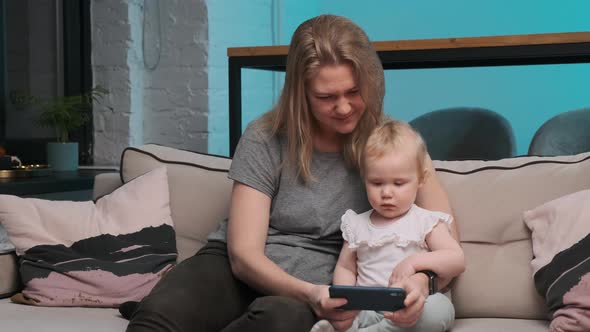 Beautiful Mother in Gray Shirt and Little Cute Girl in White Shirt Are Sitting on Couch and Looking alt