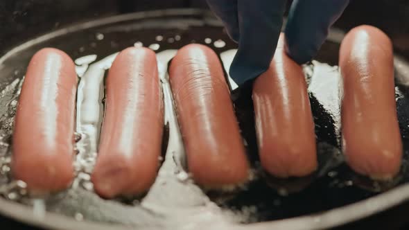 a Hand in a Blue Glove Rearranging Sausages in a Hot Frying Pan Covered with Sunflower Oil alt