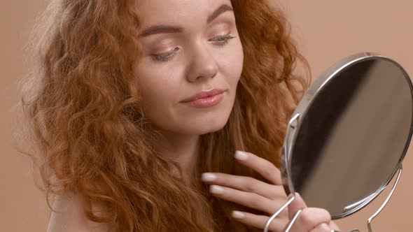 GingerHaired Lady Looking In Mirror Touching Hair On Beige Background alt