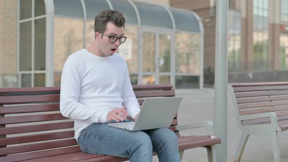 Young Man Reacting to Loss on Laptop While Sitting Outdoor on Bench alt