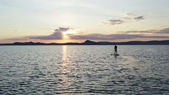A Young Woman with Long Hair is Swimming on a Board with a Paddle at Sunset