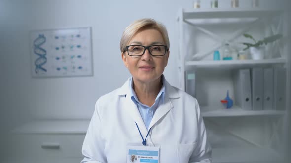 Kind Female Physician in Eyeglasses Sitting at Table and Smiling on Camera Staff