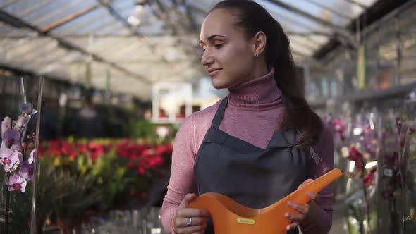 Young Female Gardener in Uniform Walking in a Greenhouse Among Shelves with Different Plants and alt
