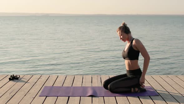Woman Practicing Yoga Sitting on Knees on Pier Tilting To Mat Ahead of Herself