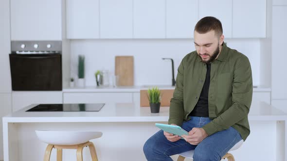 Concentrated Positive Man Sitting in Kitchen Using Tablet and Looking at Camera Smiling alt