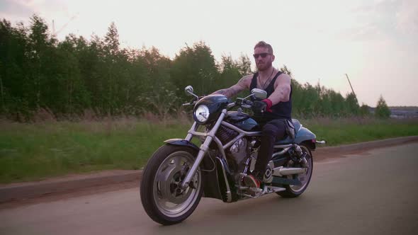 Stylish Biker with Tattoos Rides a Motorcycle on a Country Road at Sunset