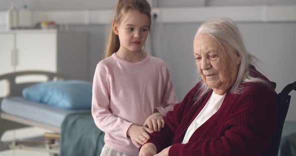 Adorable Little Girl Comforting and Supporting Old Disabled Grandmother in Hospital Ward alt
