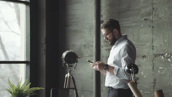 Man Withbeard Stands in Front of the Table and Uses a Smartphone alt