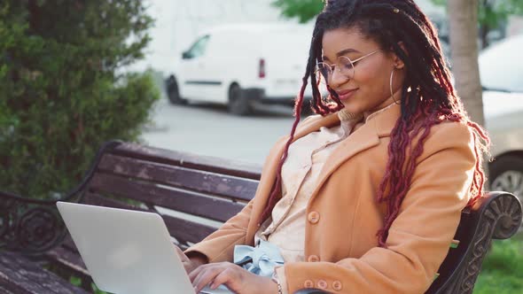 African American Woman Sitting on a Bench in City and Using Laptop