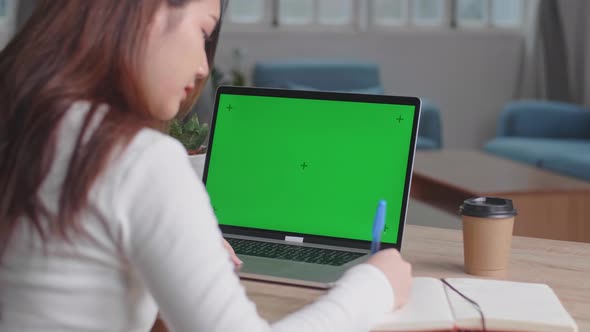 Girl Uses Laptop With Green Mock-Up Screen While Writing On Notebook In Her Living Room alt