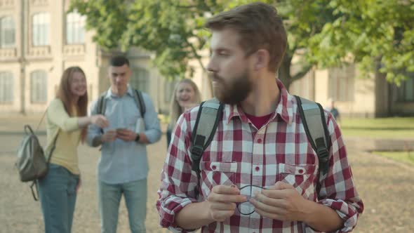 Nervous Bearded University Student Looking Back at Groupmates Laughing and Pointing at Him, and alt