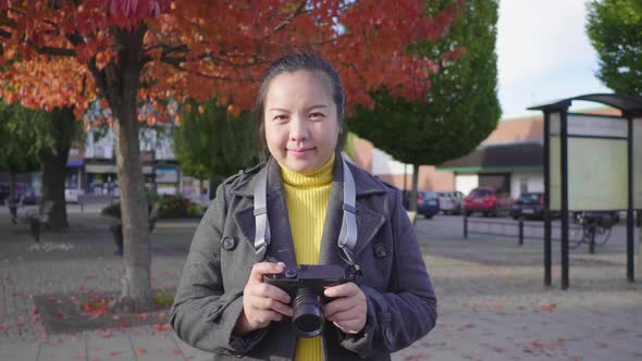 Front view of happy Asian woman standing, holding camera and smiling on the street alt