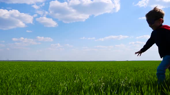 A Happy Child in a Superhero Costume in a Red Cloak Runs Across the Green Field From Left To Right alt