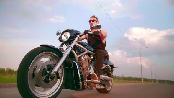 Stylish Biker with Tattoos Rides a Motorcycle on a Country Road at Sunset