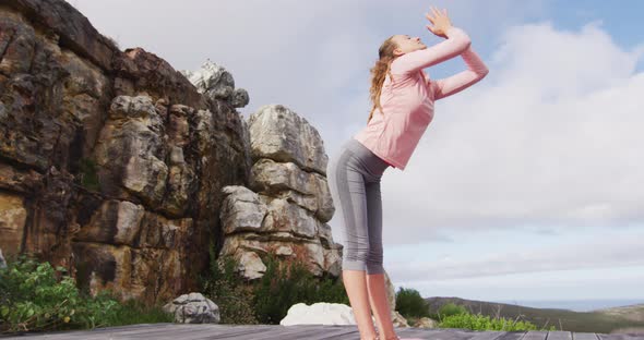 Caucasian woman practicing yoga, stretching and touching floor, standing on deck on mountainside ...