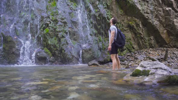 Young man walking in the stream looks at the waterfall. alt