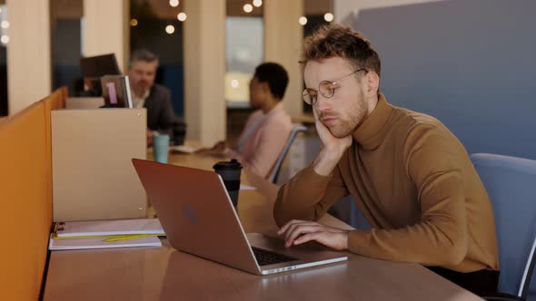 Tired Caucasian Young Man Sitting at Table and Feeling Bored While Working Hardly alt