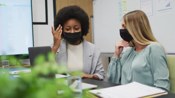 Two diverse businesswomen wearing face masks working together using laptop at desk in office alt