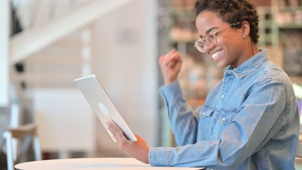 African Woman Celebrating Success on Tablet at Cafe  alt