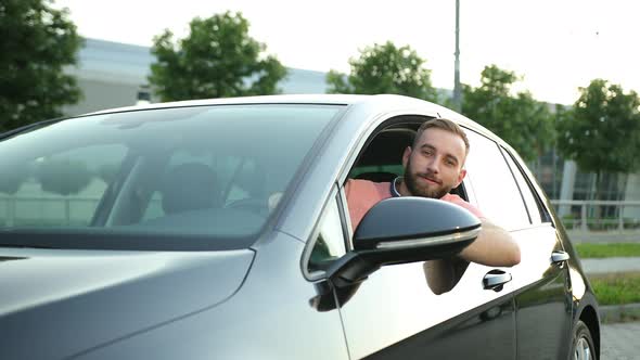 A Young Man with Beard Looks Out of the Car Window Smiles and Gives Thumbs Up alt