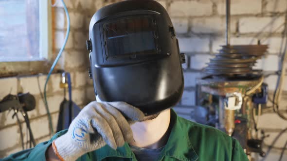 Tired Young Welder in Uniform Opens Protective Mask After Finishing His Job alt