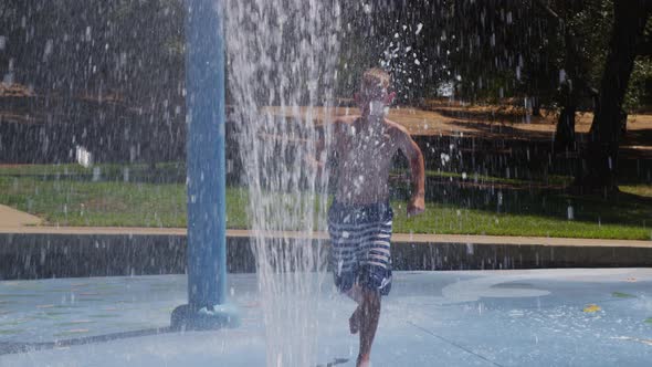 Boy jumping through water fountain on summer day, slow motion alt