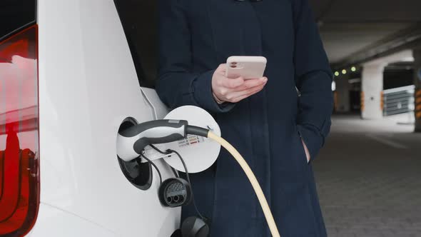 Woman Standing Near Charging an Electric Car and Using Her Mobile Phone alt
