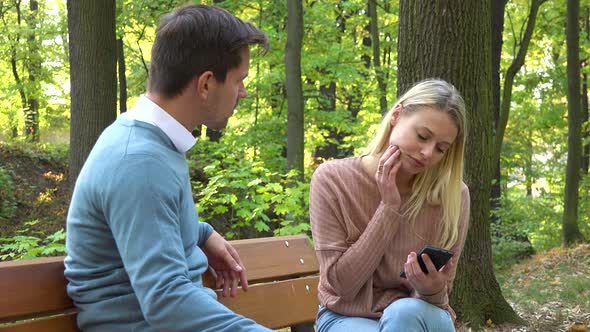 A Man and a Woman Sit in a Park, the Woman Is Absorbed with Her Smartphone, the Man Is Angry at Her alt