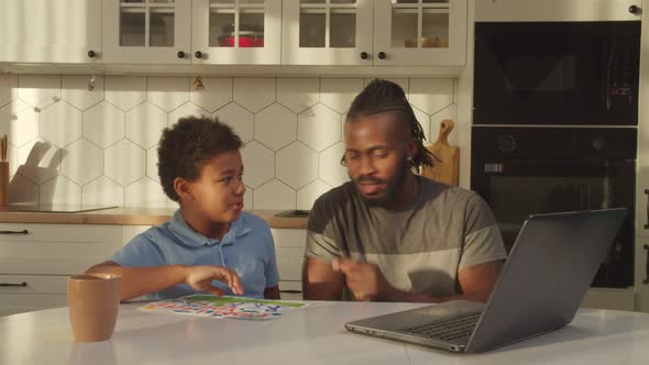 Joyful African Father and Cute Boy Making Video Call Online Using Laptop Indoors alt