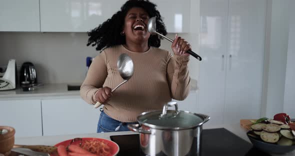 Black young african woman having fun cooking and dancing inside kitchen at home alt
