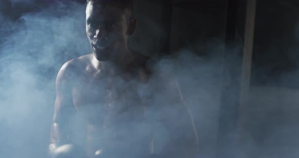 African american man wearing boxing gloves banging fists together in dark smoky room alt