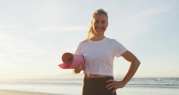 Portrait of caucasian woman holding yoga mat at the beach smiling alt