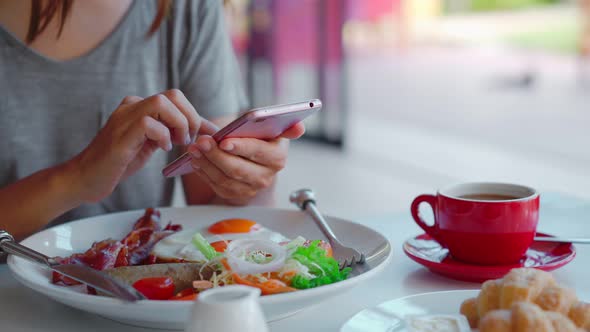 Young woman using smart phone with cup of coffee while eating breakfast. alt