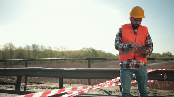 Young Bearded Man Inspector in an Orange Vest and Protective Helmet with Tablet alt