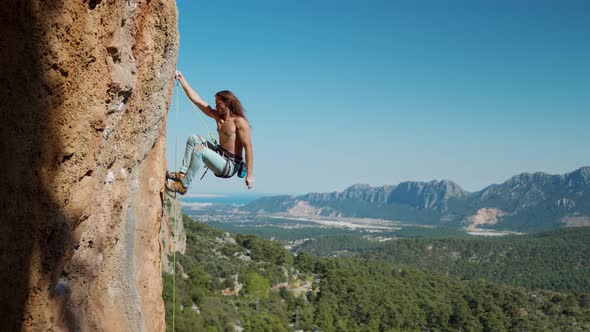 Attractive Strong and Fit Man Rock Climber with Long Hair Hangs on Rope on Vertical Cliff and Looks alt