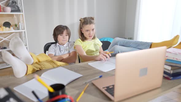 Wide Shot of Relaxed Caucasian Children with Feet on Table Looking at Laptop and Talking alt
