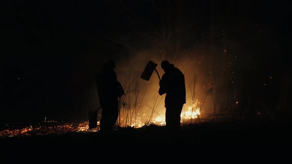 Two Firefighters with Fire Flappers Extinguish a Fire at Night alt