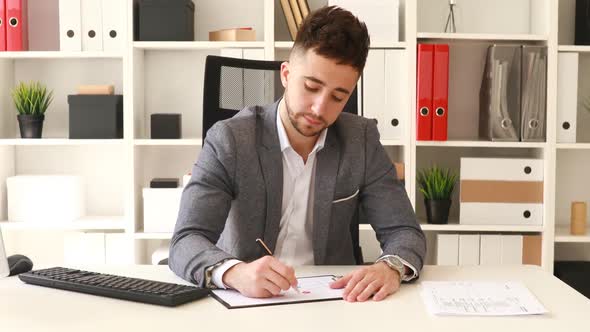 Businessman in Gray Jacket Sitting at Table in White Office and Writing on Documents