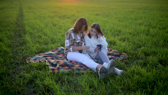 Young mum with a cute daughter use smartphone sit on plaid on a field at sunset
