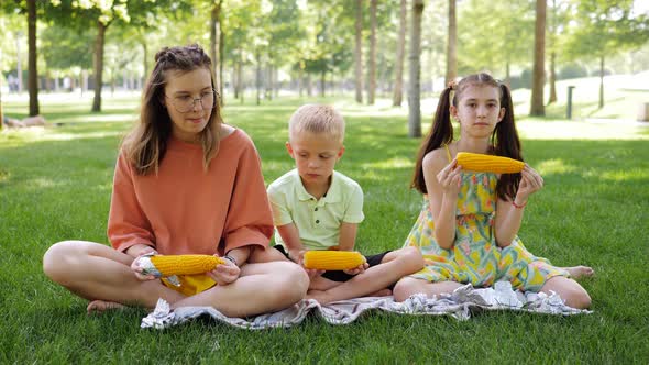 Three Cute Children Boy and Two Girls of Different Ages Eating Boiled Corn Cobs During Picnic in the