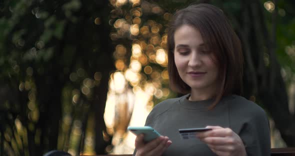 Student on Street with Credit Card and Phone Brunette Woman Sitting on the Bench and Makes Online alt