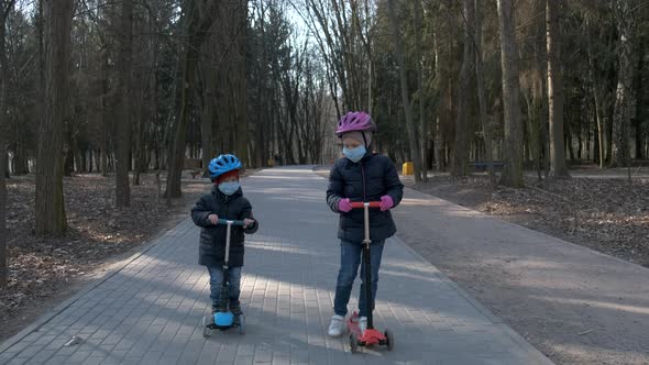 Little Boy and Girl Ride Scooters in Medical Masks in the Park. The Concept of Protection FROM Covid