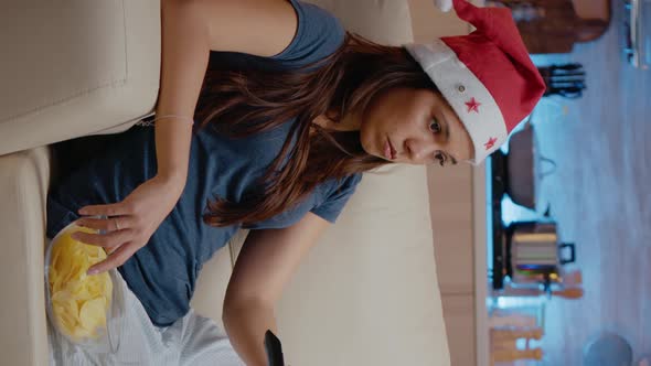 Vertical Video Woman with Santa Hat Switching Channels Using TV Remote Control alt