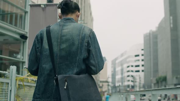 Handsome Asian man wearing a mask using smartphone and carrying shoulder bag walking.